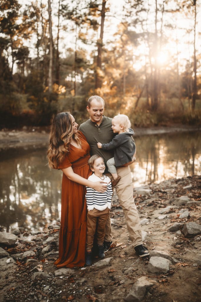 Family Photographer - A husband, wife and their two sons stand near quiet river behind them